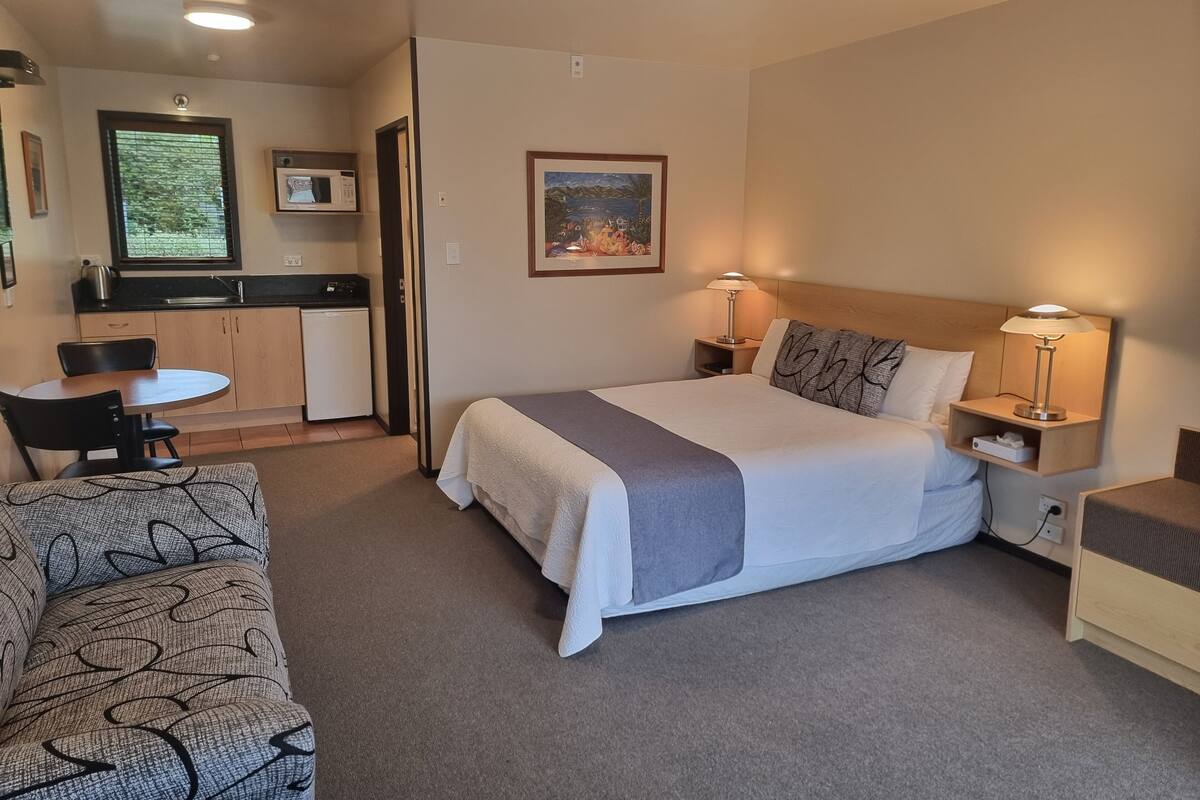A hotel room with a bed, bedside tables, a desk, a chair, a small kitchenette, and a sofa.