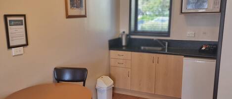 Una pequeña kitchenette con microondas, un lavamanos y una ventana. Hay un cubo de basura y un cuadro en la pared.