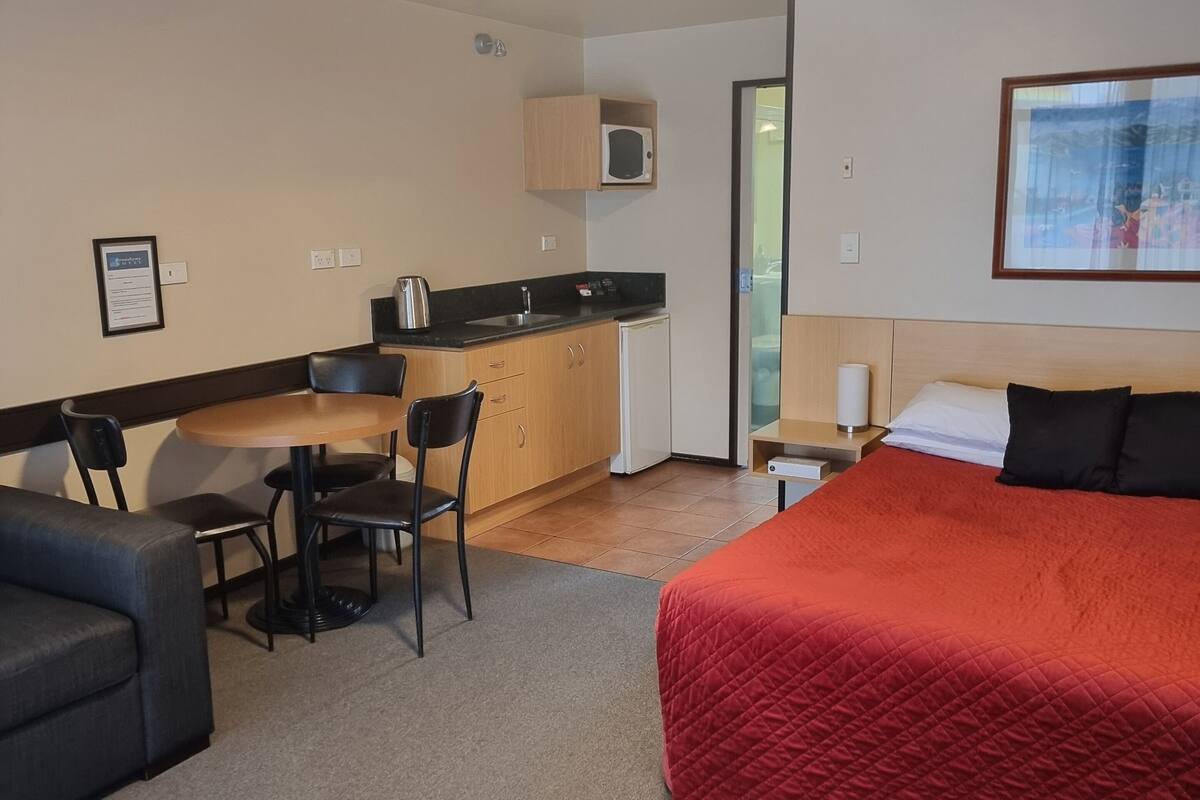 A hotel room with a bed, a desk, a chair, a kitchenette, and a framed picture on the wall.