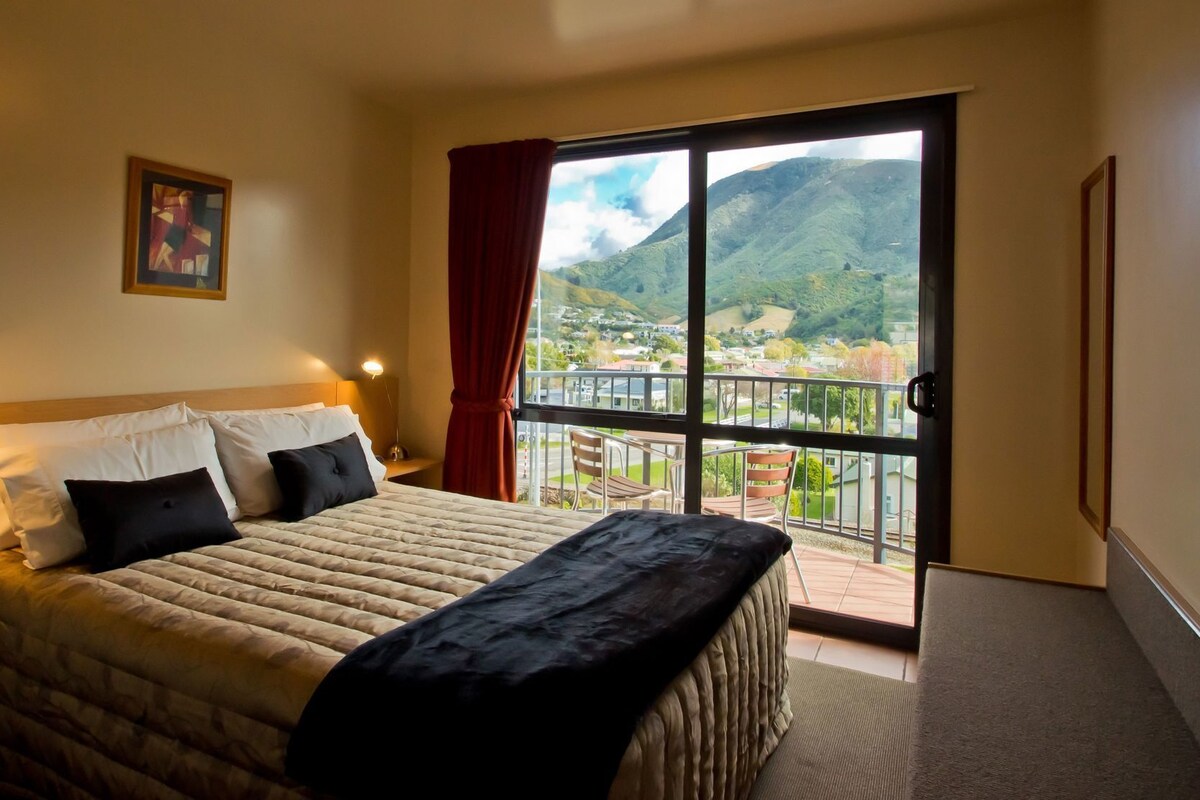 A hotel room with a bed, a nightstand, a window with a view of a mountainous landscape, and a door to a balcony.
