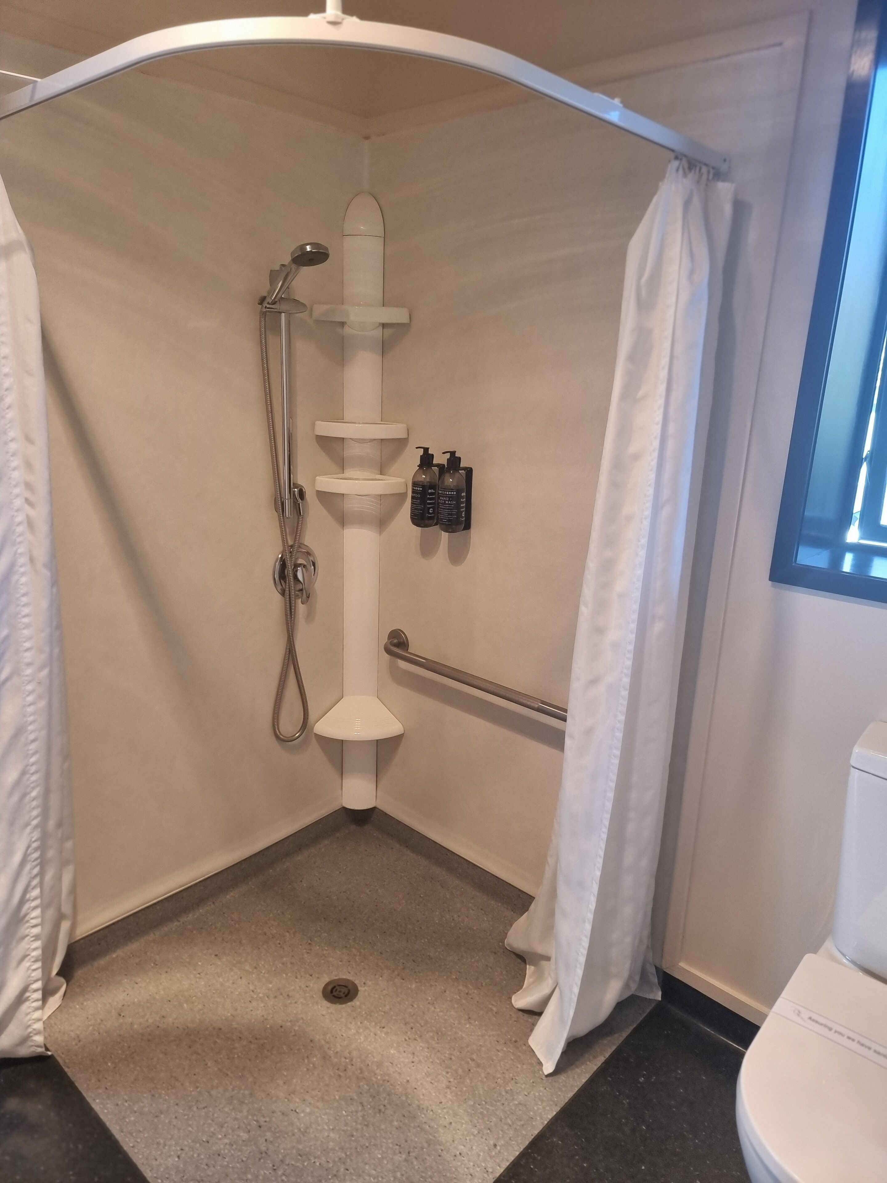 A small, enclosed shower area with a grab bar, a handheld showerhead, and two bottles of shampoo.