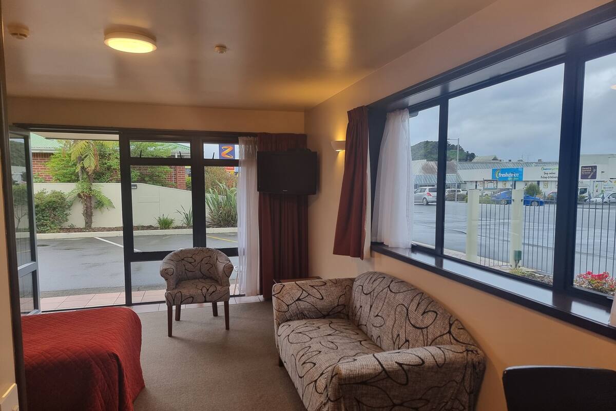 A hotel room with a large window, a sofa, a chair, and a bed with red bedding.