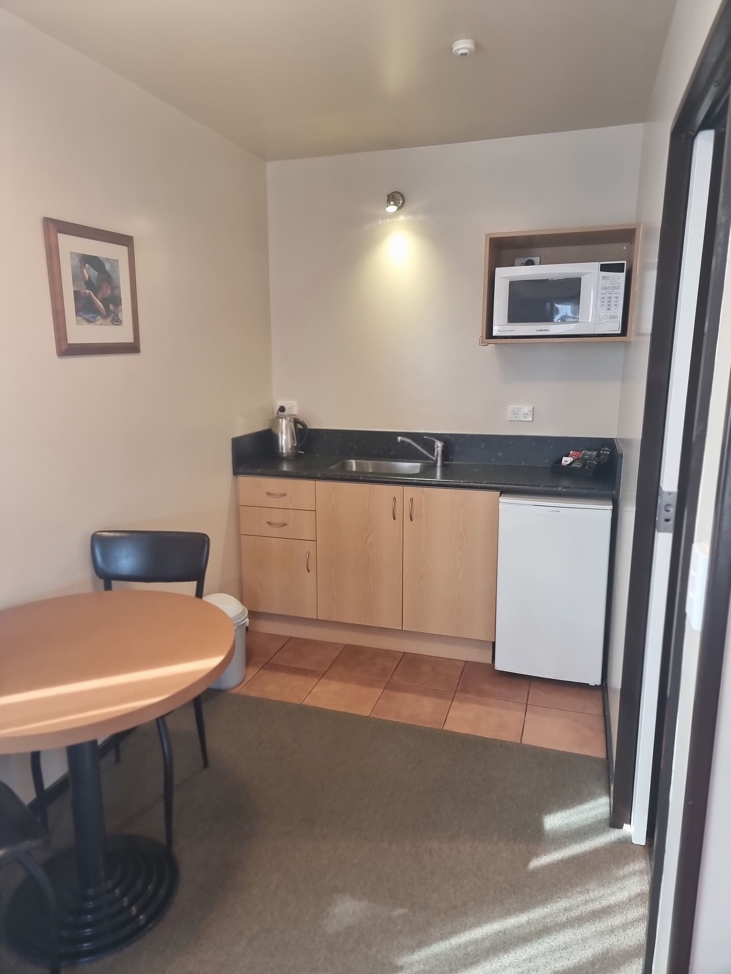 Een compacte keuken met een magnetron, spoelbak en een kleine koelkast. Er is een stoel en een tafel, en een fotolijst aan de muur.