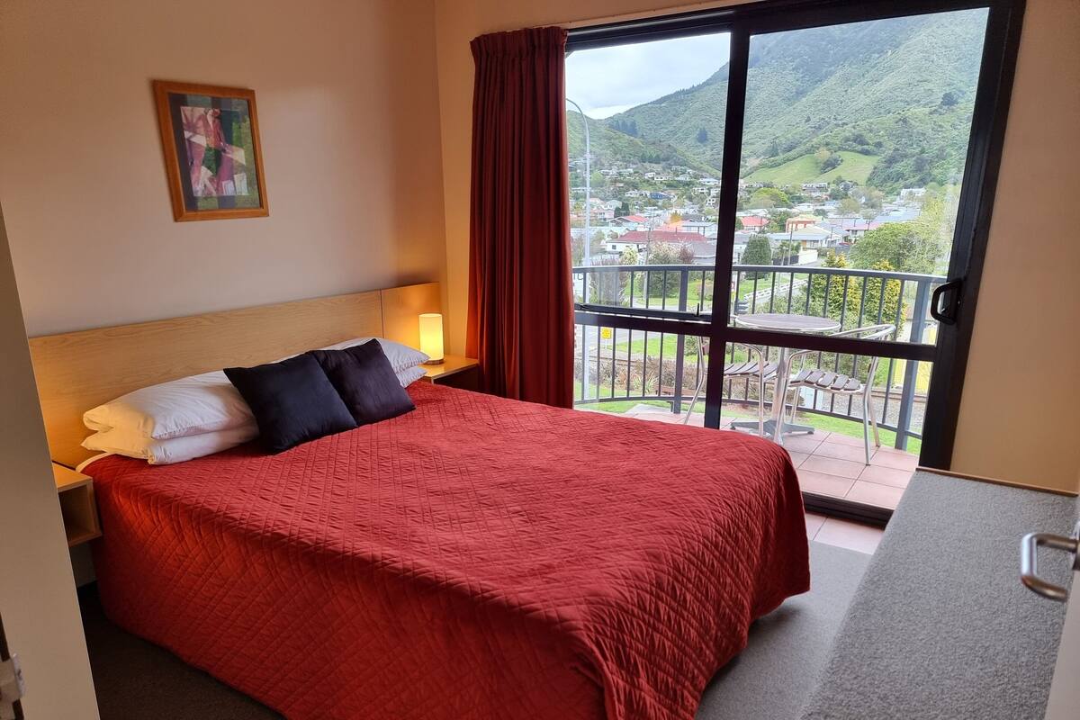 A hotel room with a bed, a nightstand, a painting, and a balcony with a view of a town and mountains.