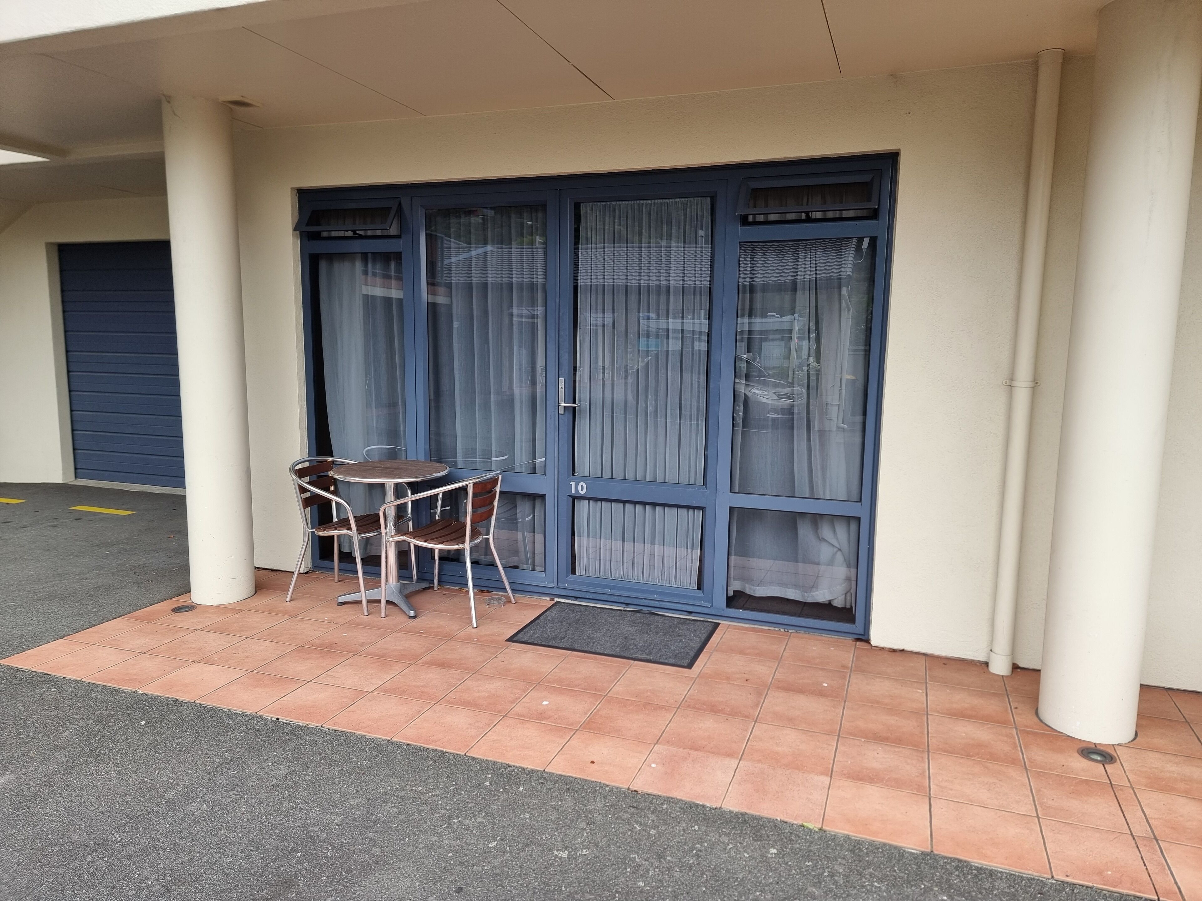 A covered outdoor seating area with a table and chairs, a blue door with a number, and a blue garage door in the background.