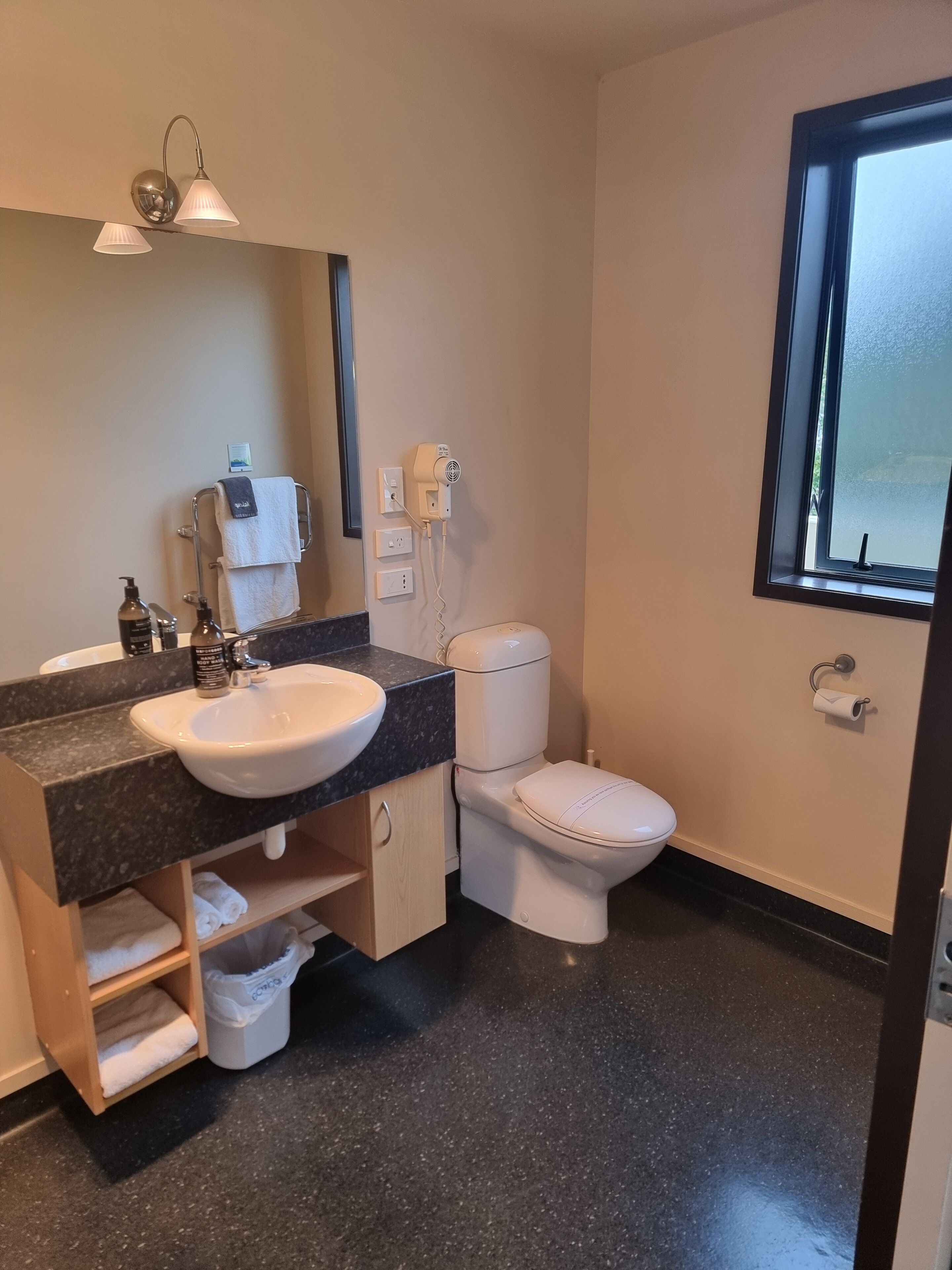Ein modernes Badezimmer mit einem weißen Waschbecken, einer dunklen Arbeitsplatte und einer Toilette. Ein Fenster bietet einen Blick nach draußen, und ein Handtuchhalter trägt ein Handtuch.