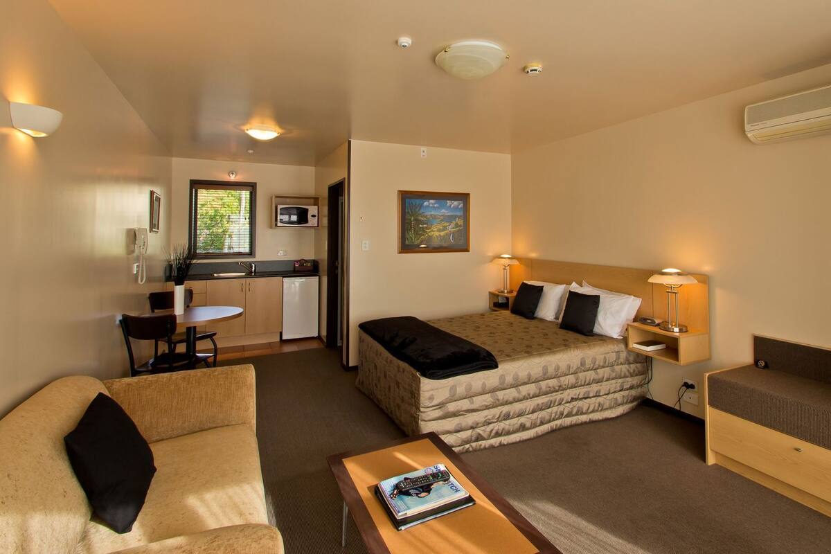 A compact hotel room with a bed, a sofa, a small table, a desk with a microwave, and a kitchenette.