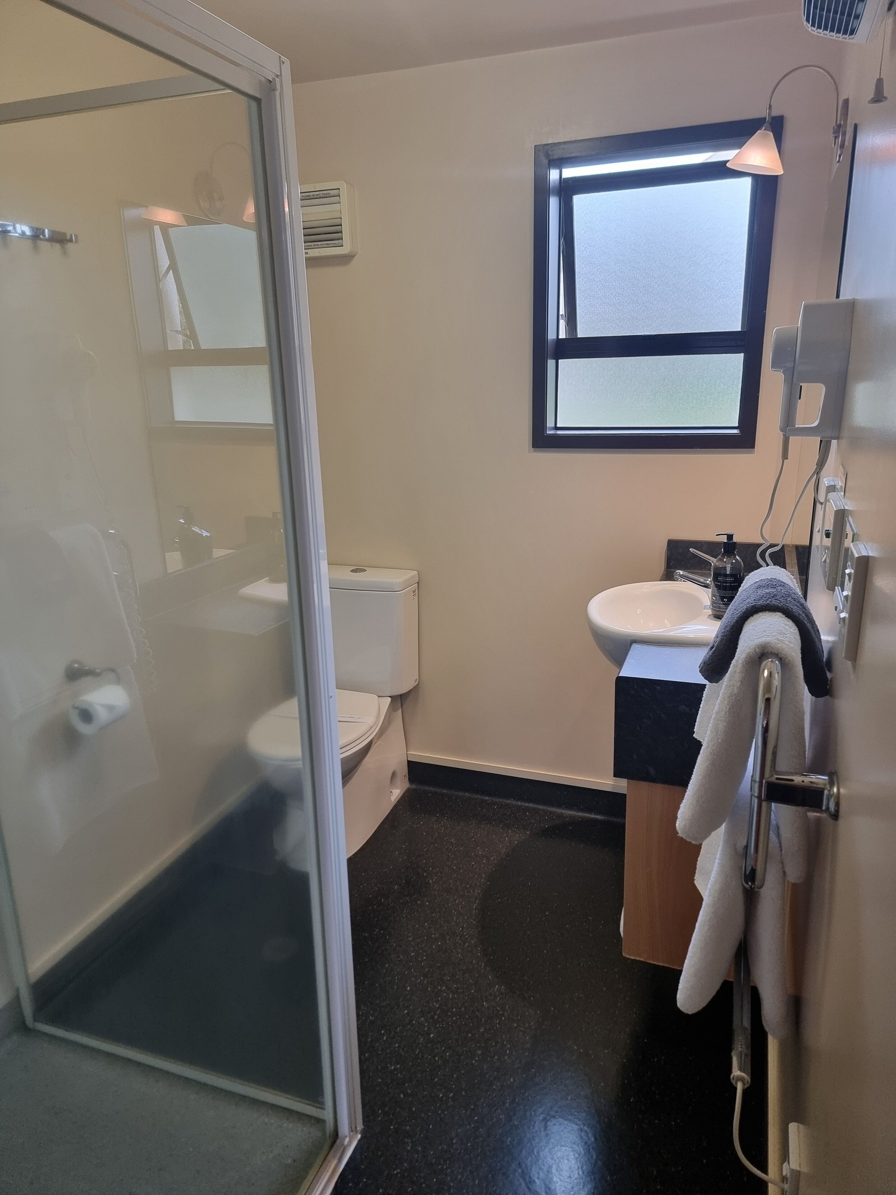 A bathroom with a glass shower door, a toilet, a sink with a towel rack, and a window providing natural light.