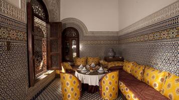 Un interior tradicional marroquí con un comedor que presenta un piso de baldosas estampadas, un sofá con cojines amarillos y una puerta de madera decorativa.