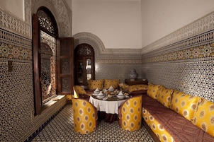 Um interior tradicional marroquino com área de refeições, chão de azulejos estampados, sofá com almofadas amarelas e porta de madeira decorativa.