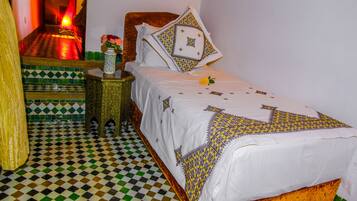 Una habitación con piso a cuadros, una cama con ropa de cama estampada, una mesita de noche con un jarrón y un calefactor en la pared.