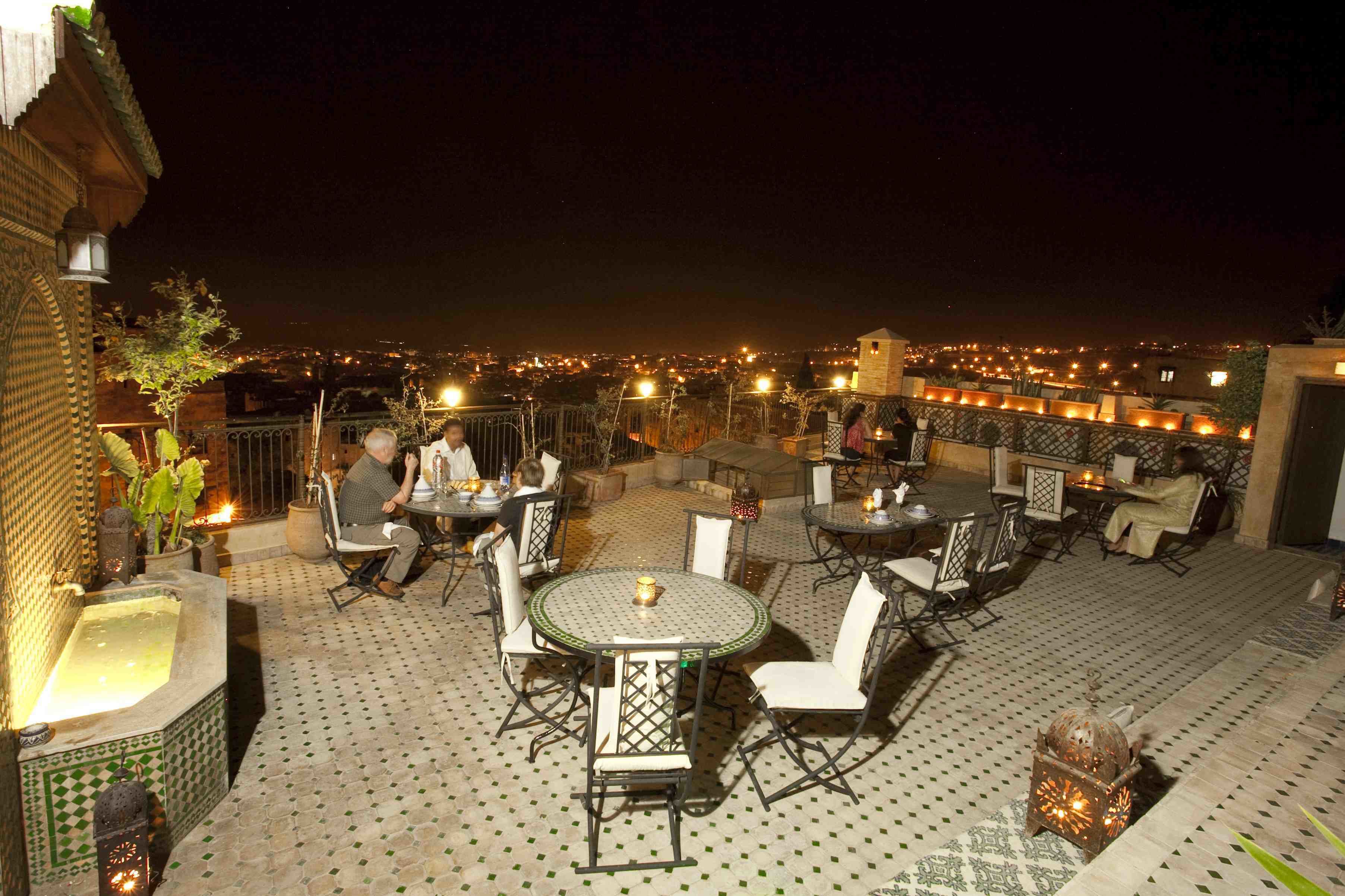 Um pátio externo com mesas e cadeiras redondas, uma fonte decorativa e vista da cidade à noite.