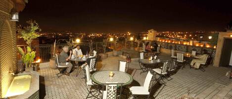 An outdoor patio with round tables and chairs, a decorative fountain, and a view of the city at night.