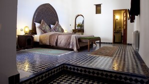 Une chambre avec un sol à carreaux, un grand lit avec une tête de lit, une table de chevet avec une lampe et un miroir au mur.