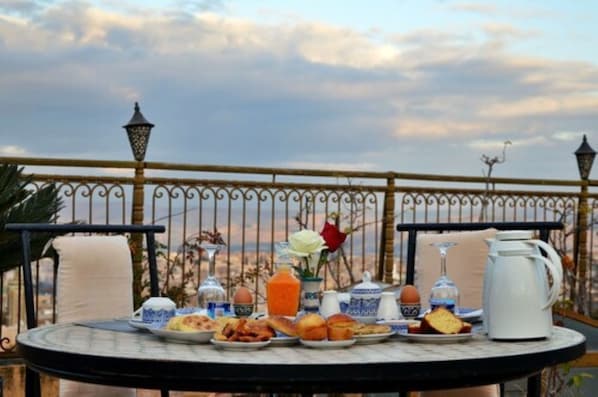Una mesa dispuesta con alimentos para el desayuno, como pasteles, huevos y bebidas, en un balcón con vistas al horizonte de la ciudad.