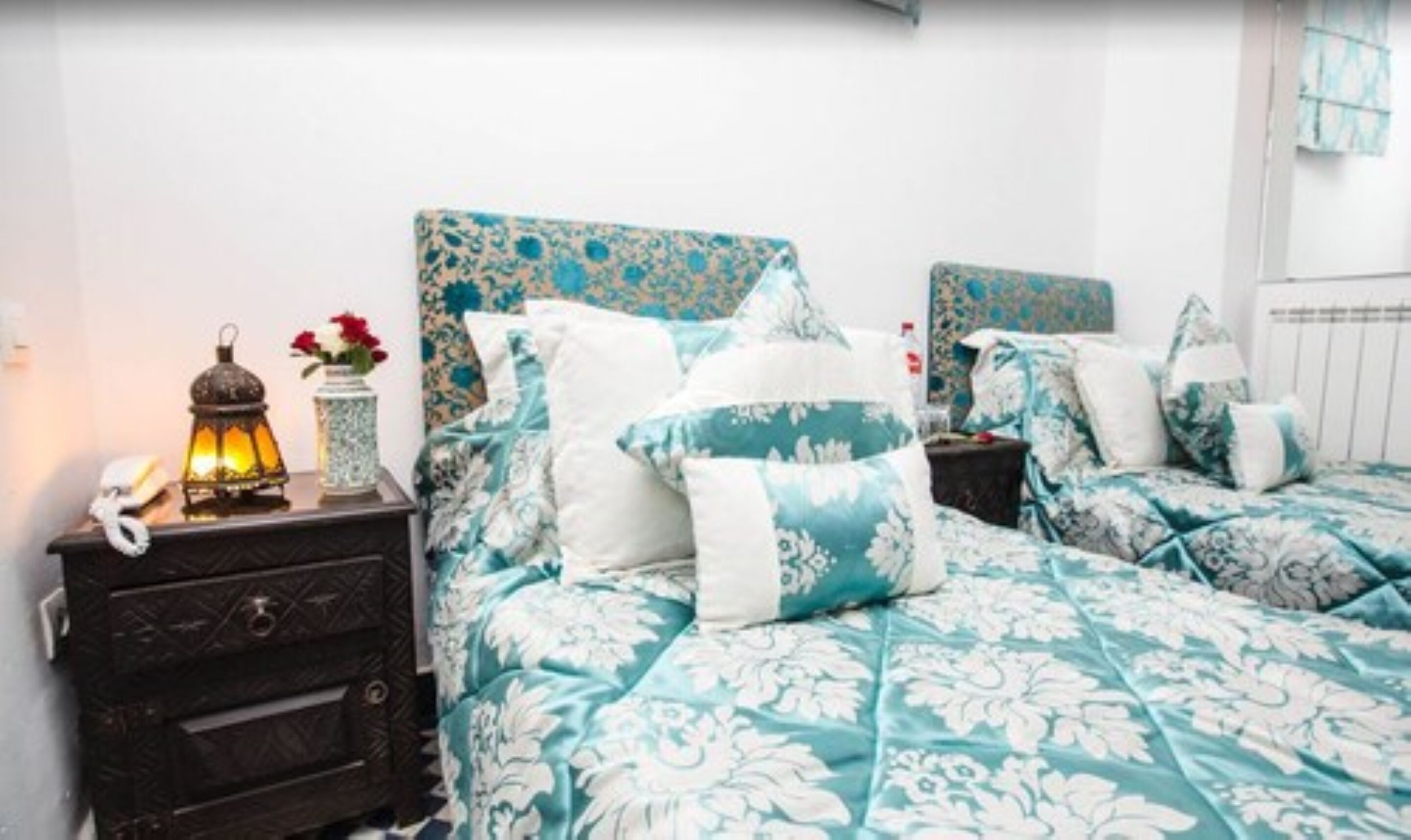 Une chambre avec deux lits, une literie à motifs floraux, une table de chevet avec une lampe et un vase rempli de fleurs.