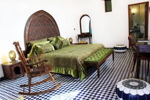 Quarto com piso xadrez, uma cadeira de balanço de madeira, uma cama estofada verde e um espelho na parede.