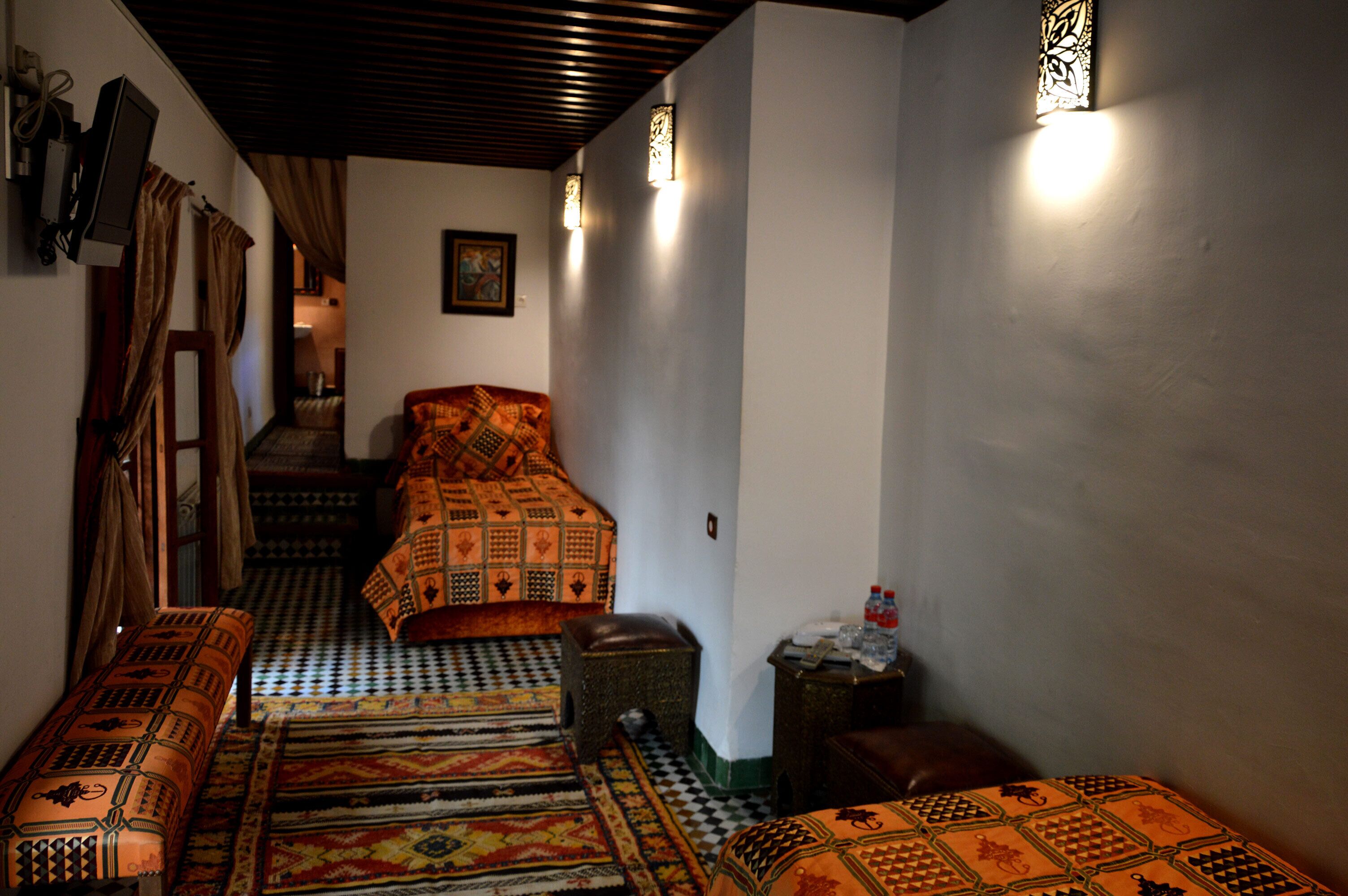 Un couloir étroit, avec un lit au fond, un banc, des tapis à motifs et des lampes fixées au mur.