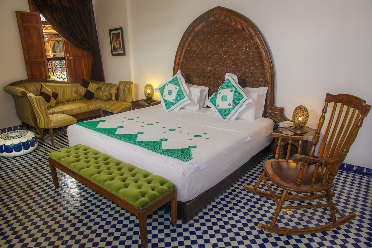 Une chambre à coucher bien aménagée, avec un sol à carreaux, un grand lit recouvert d’une couette verte et blanche, un banc vert à motifs capitonnés, un fauteuil à bascule en bois et un canapé agrémenté de coussins à motifs.