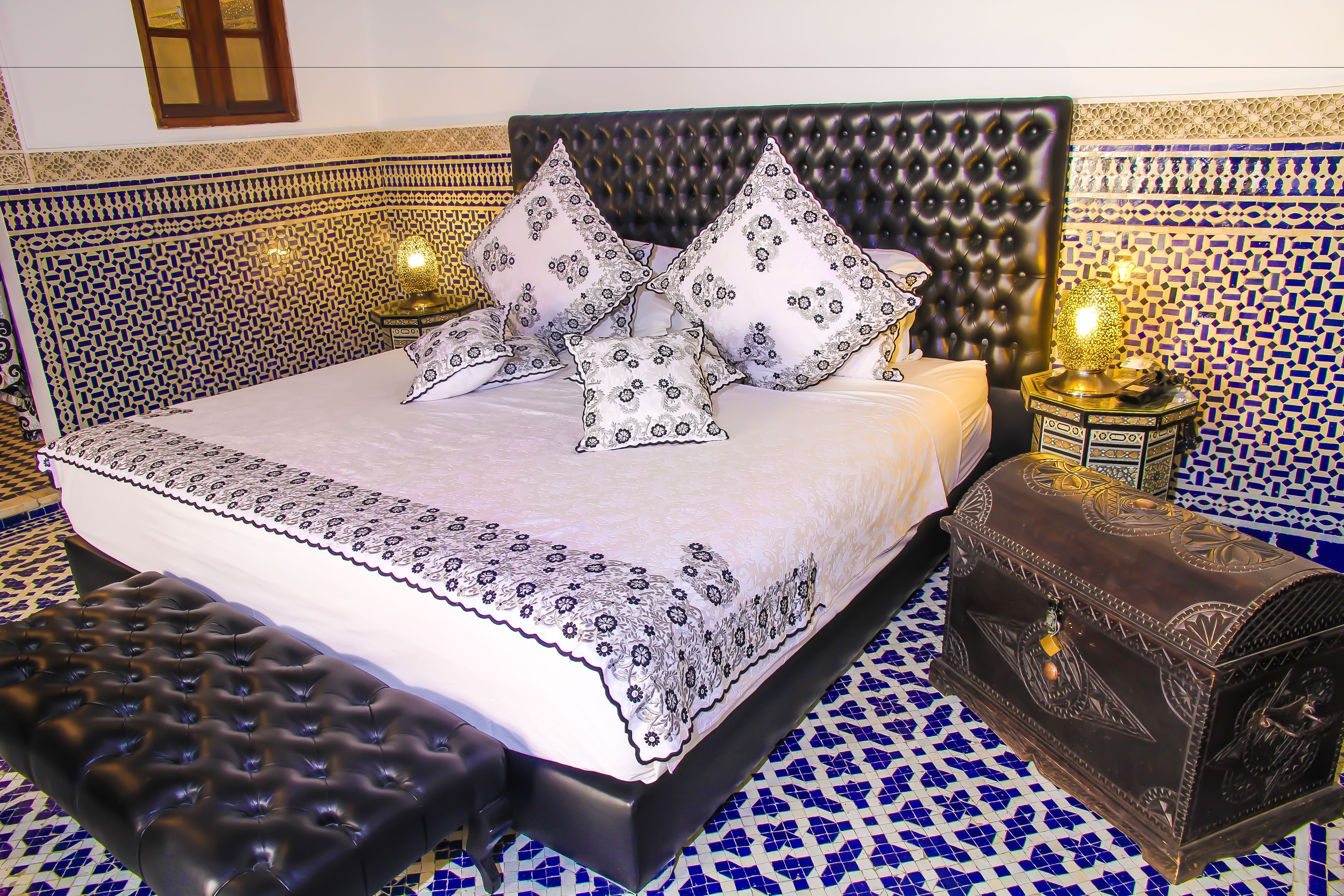 Una cama con colcha estampada y varias almohadas, cabecero capitonado, cómoda decorativa y suelo con diseño.