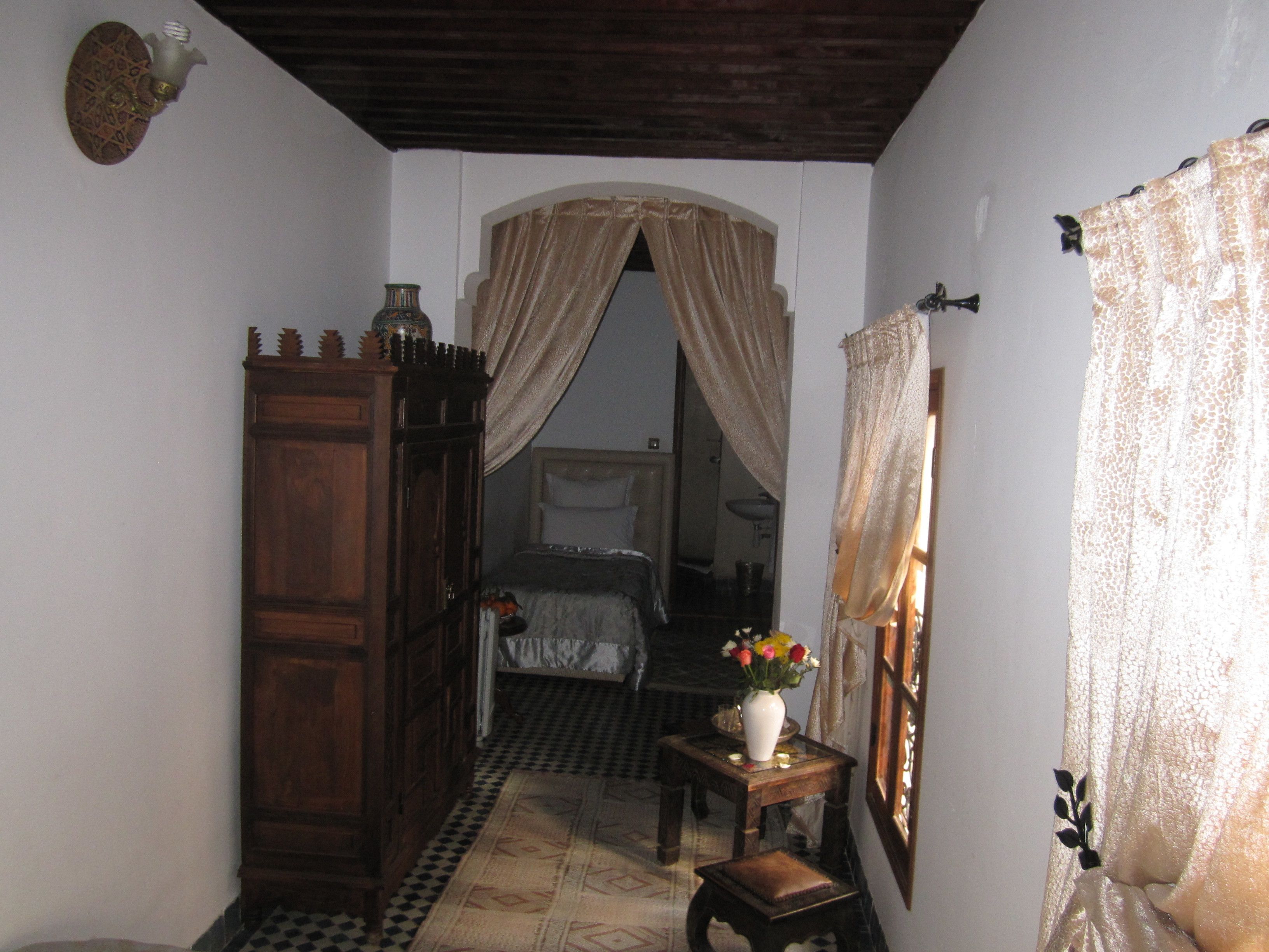 A narrow hallway with a wooden wardrobe, a small table with a vase of flowers, and a bed with a canopy.