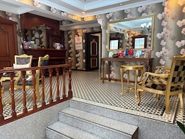 A hotel lobby with a checkered floor, a staircase, wooden furniture, and a floral wallpaper design.