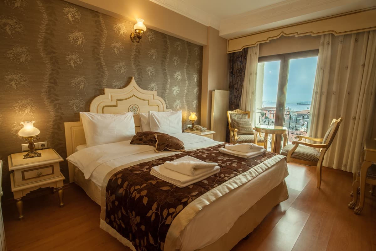 A hotel room with a large bed, two bedside tables, a chair, a small table, and a view of the sea.