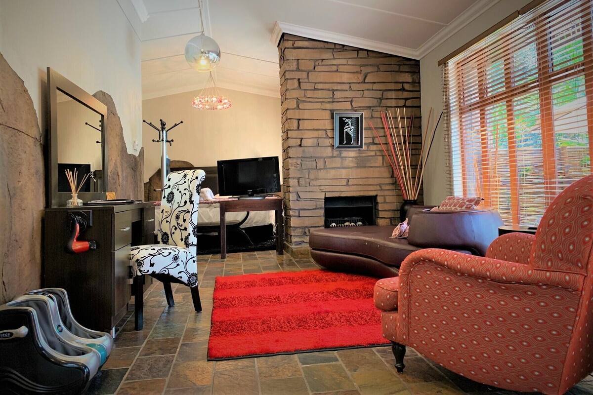 A living room with a stone fireplace, a television, a patterned armchair, a red rug, and a large window with blinds.