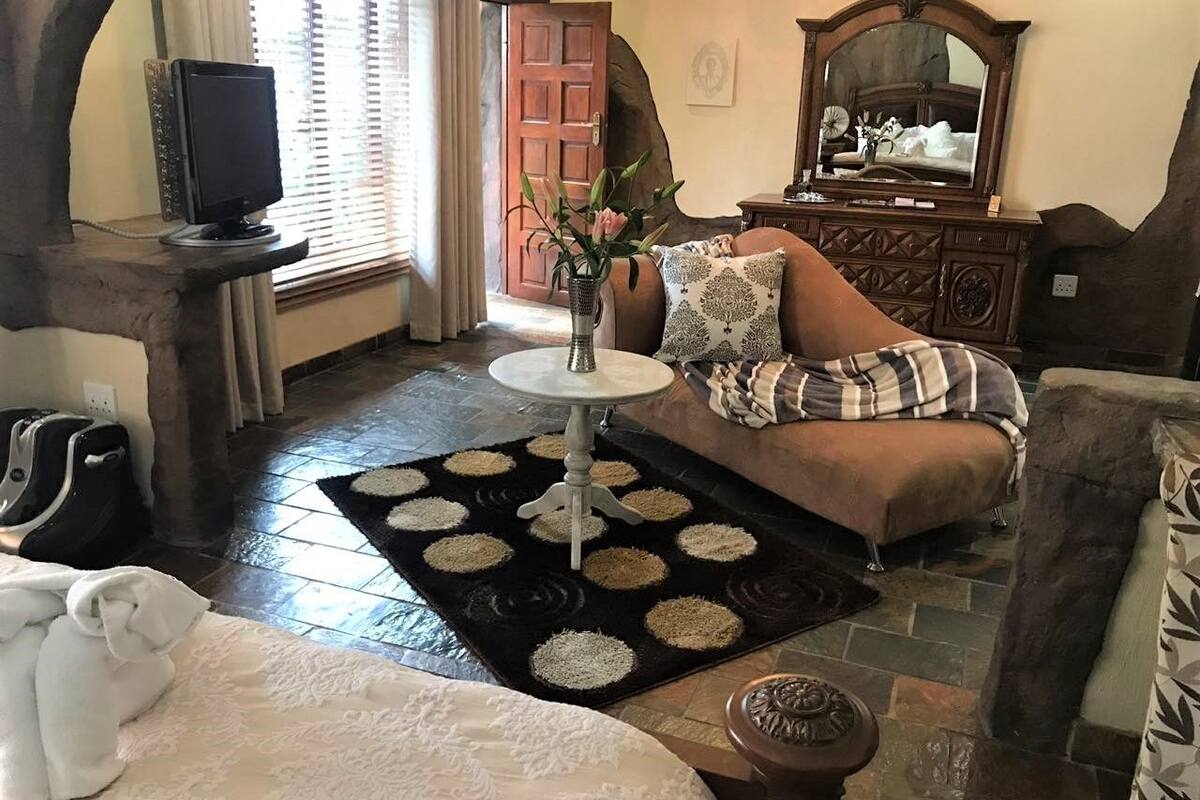 A cozy living room with a stone fireplace, a round table with a vase, a sofa with patterned cushions, and a large mirror above a wooden dresser.