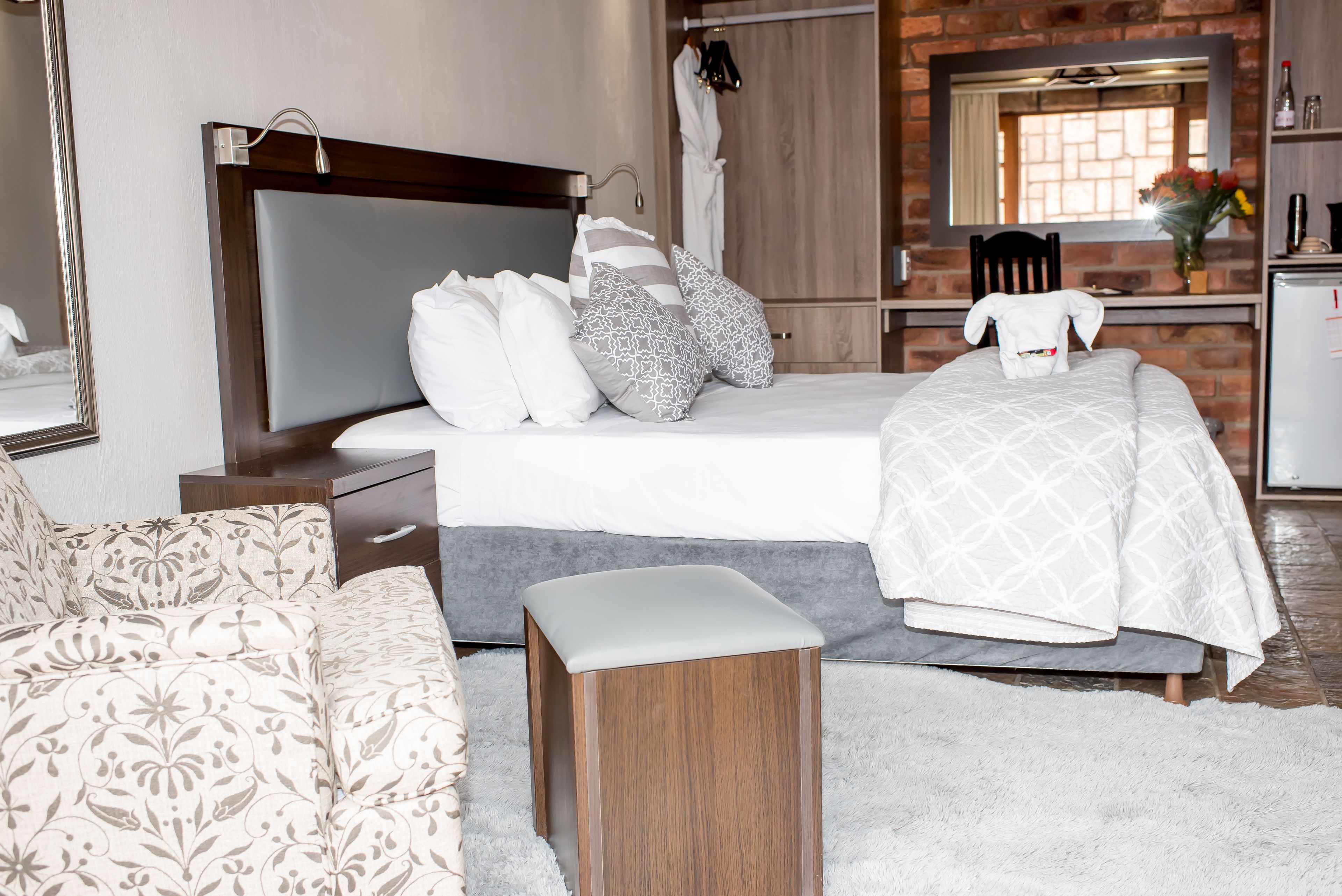 A modern hotel room with a large bed, a wooden nightstand, a small stool, a white sofa, and a bathroom visible through an open door.
