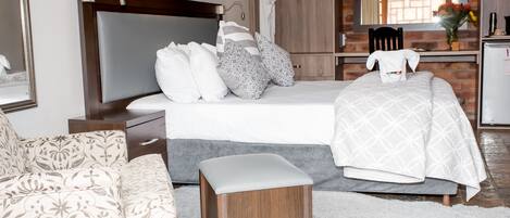 A modern hotel room with a large bed, a wooden nightstand, a small stool, a white sofa, and a bathroom visible through an open door.