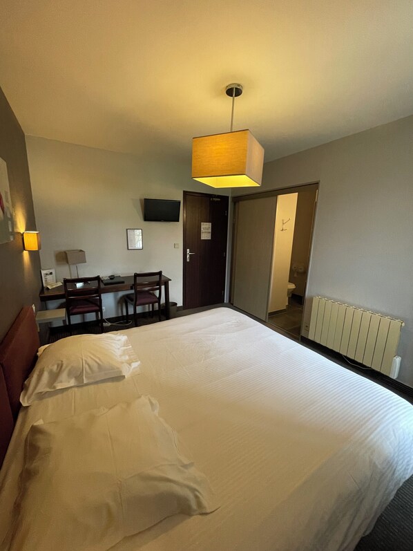 A hotel room with a large bed, a desk with a chair, a television on the wall, and a door leading to another room.
