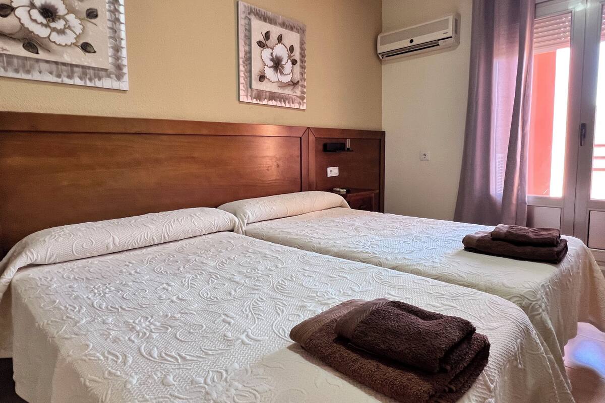 Habitación de hotel con dos camas, cabecero de madera, aire acondicionado y una ventana con cortinas.