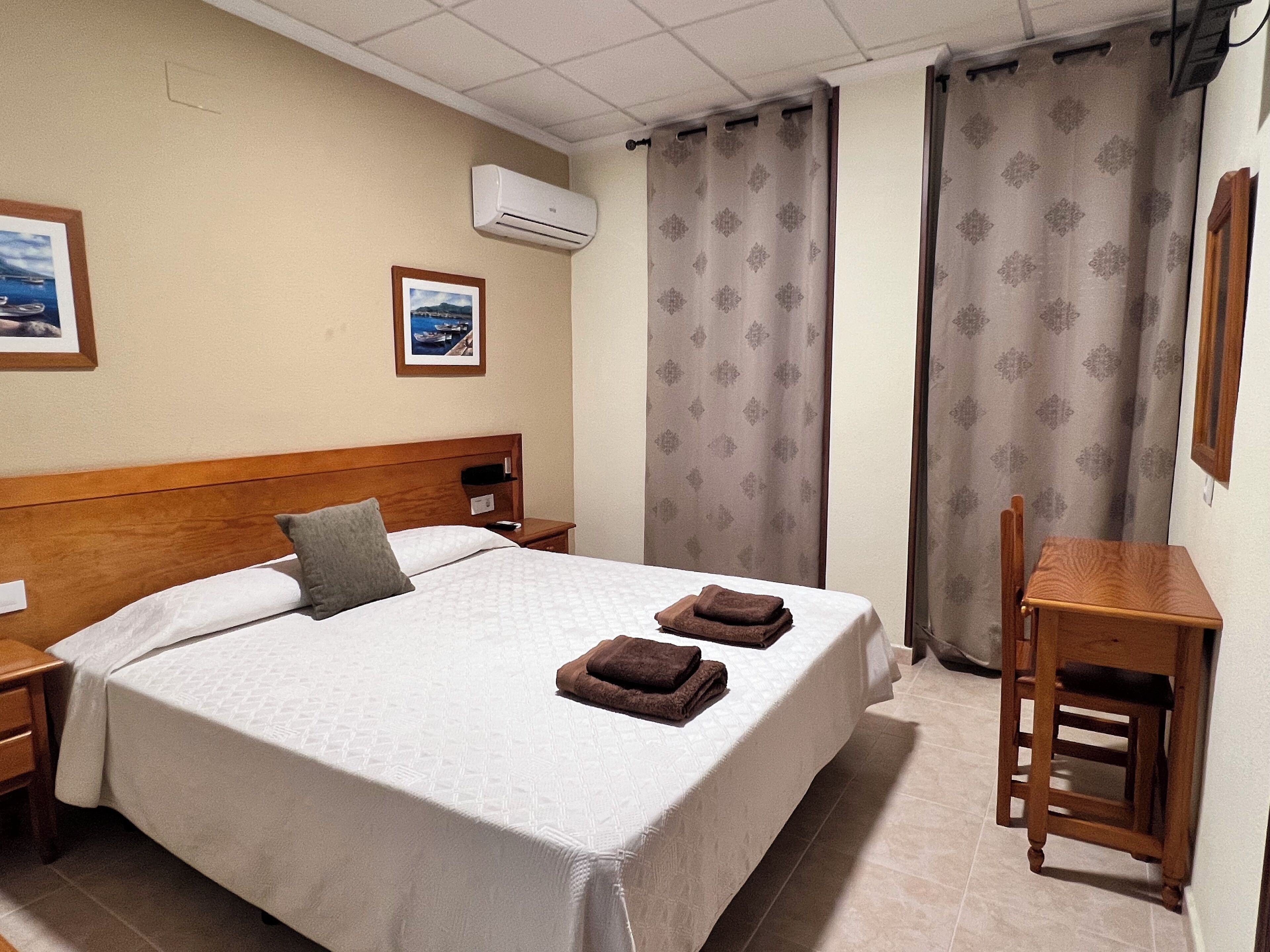 Habitación de hotel con una cama grande, cabecera de madera, mesita de noche, escritorio y una silla. Se observa un cuadro en la pared y un equipo de aire acondicionado sobre la cama.