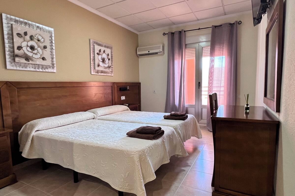 Habitación de hotel con dos camas, cabecera de madera, escritorio, una silla y ventana con cortinas.