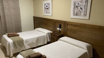 Habitación de hotel con dos camas, cada una con cubrecama blanco y toalla marrón. Hay dos cuadros de flores colgados en la pared, cabecera de madera, mesita de noche con teléfono y un equipo de aire acondicionado en la pared.