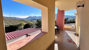 Een balkon met een rood dak, een tafel en stoelen, met uitzicht op een glooiend landschap met bergen en hoogspanningslijnen.