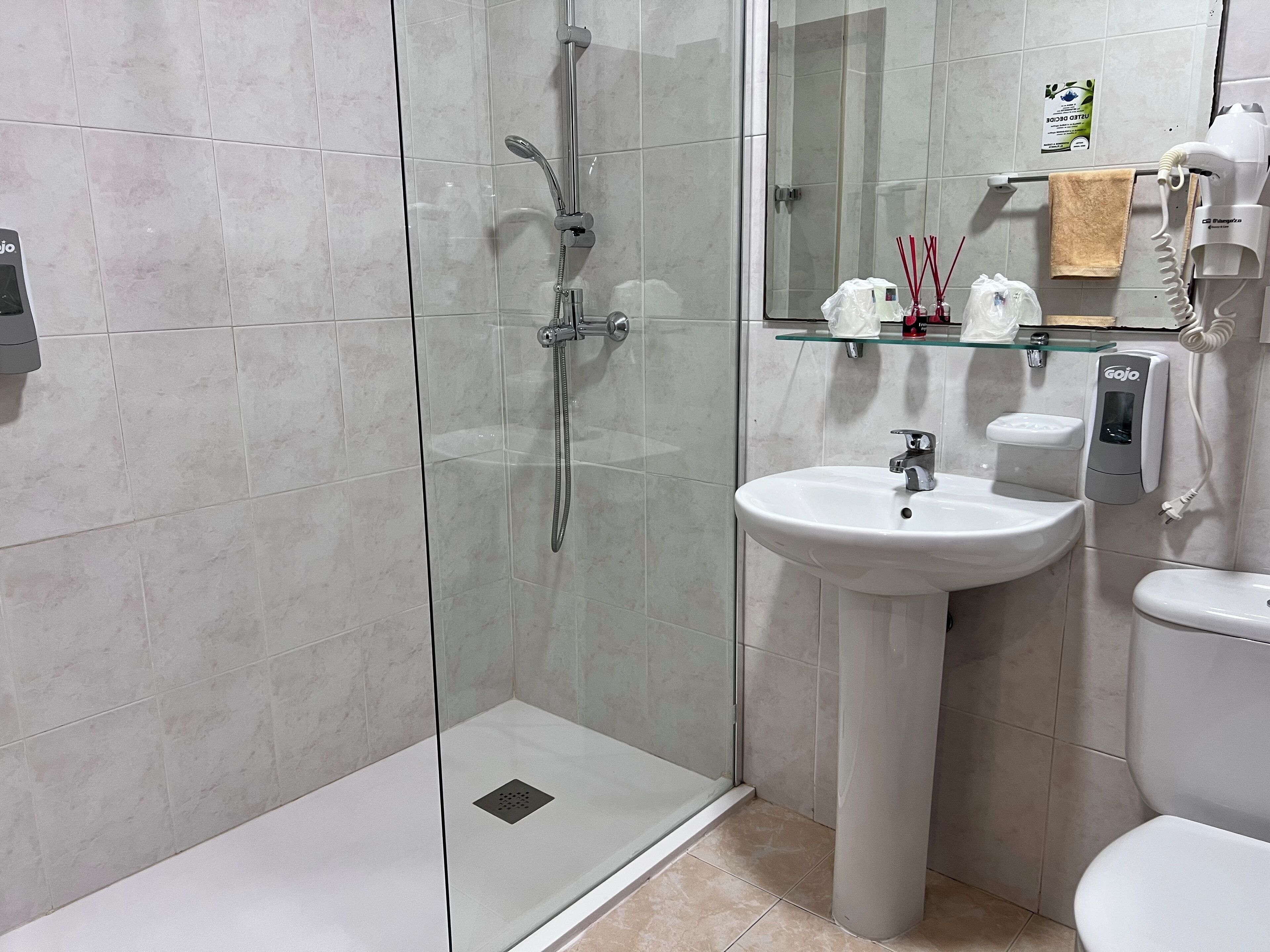 Une salle de bain moderne dotée d’une douche à parois de verre, d’un lavabo sur colonne, de toilettes et d’une étagère en verre où sont rangés les articles de toilette.
