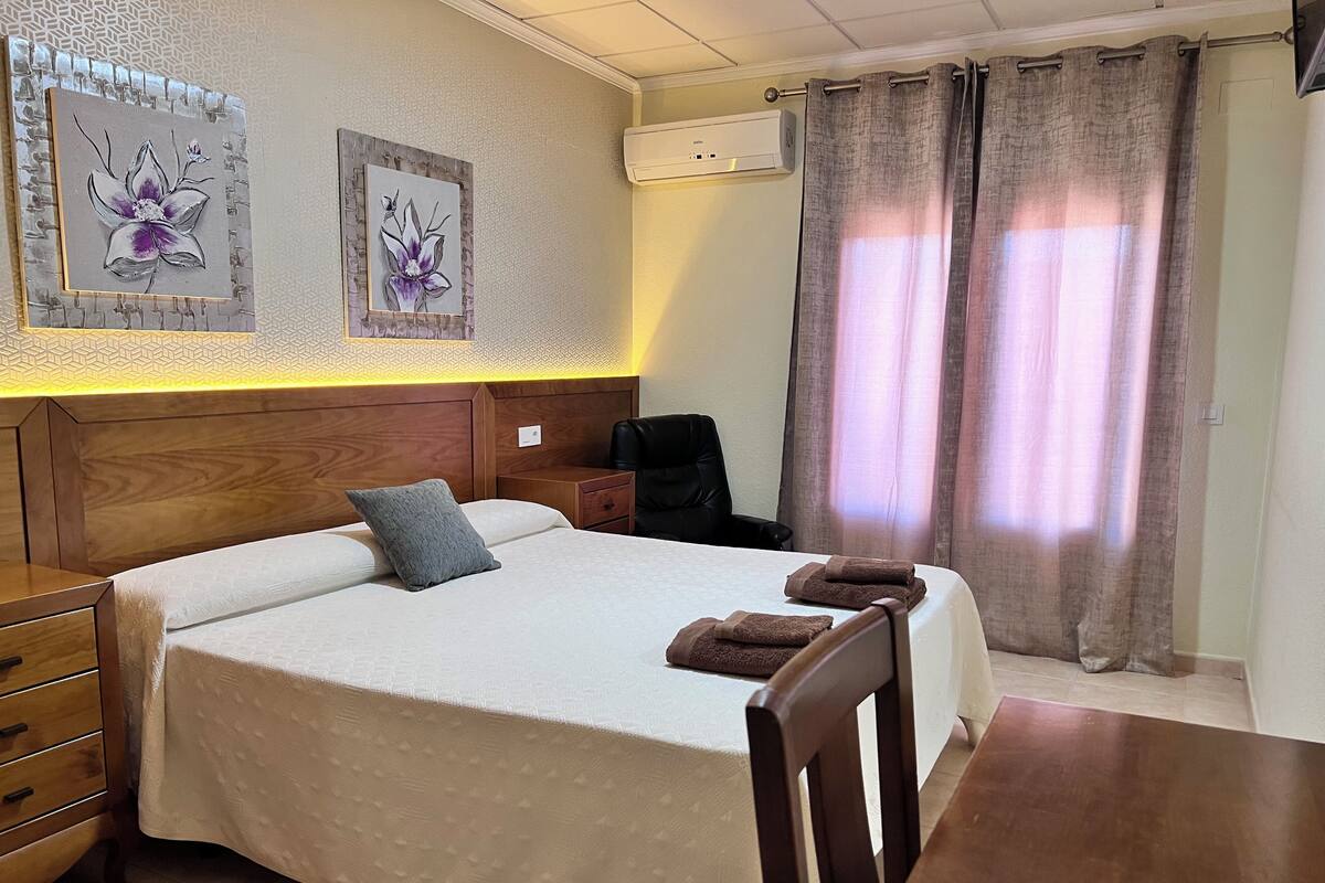 Una habitación de hotel con cabecera de madera, cama con colcha blanca, almohada gris, mesita de noche de madera, silla negra, mesa de madera y dos unidades de aire acondicionado fijadas a la pared.