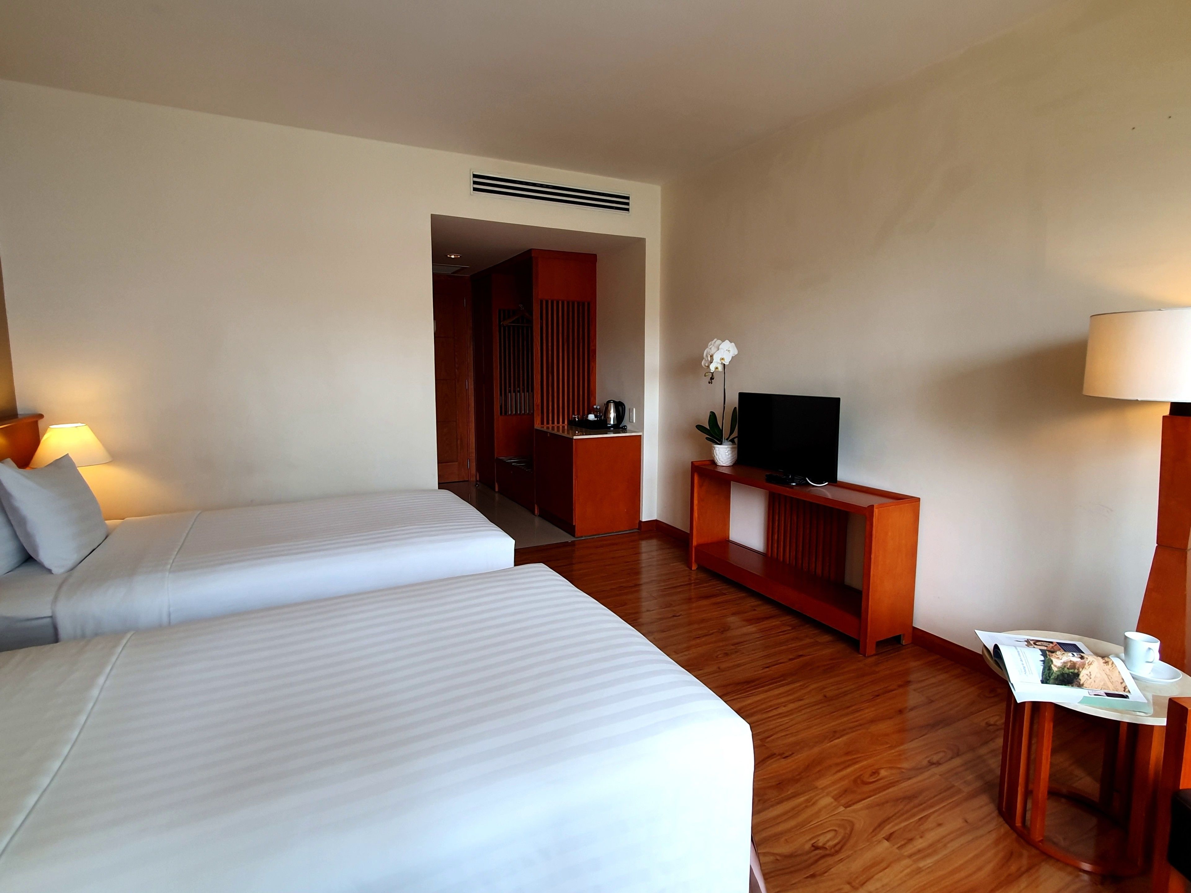 Habitación de hotel con dos camas, un televisor sobre una consola de madera, una mesita con lámpara y un pasillo que conduce a un baño.