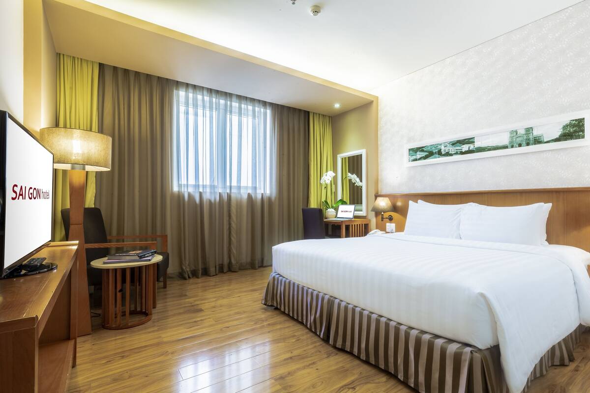 Une chambre d’hôtel avec un grand lit, un bureau avec une lampe, une chaise, une petite table avec une plante et un tableau encadré au mur.