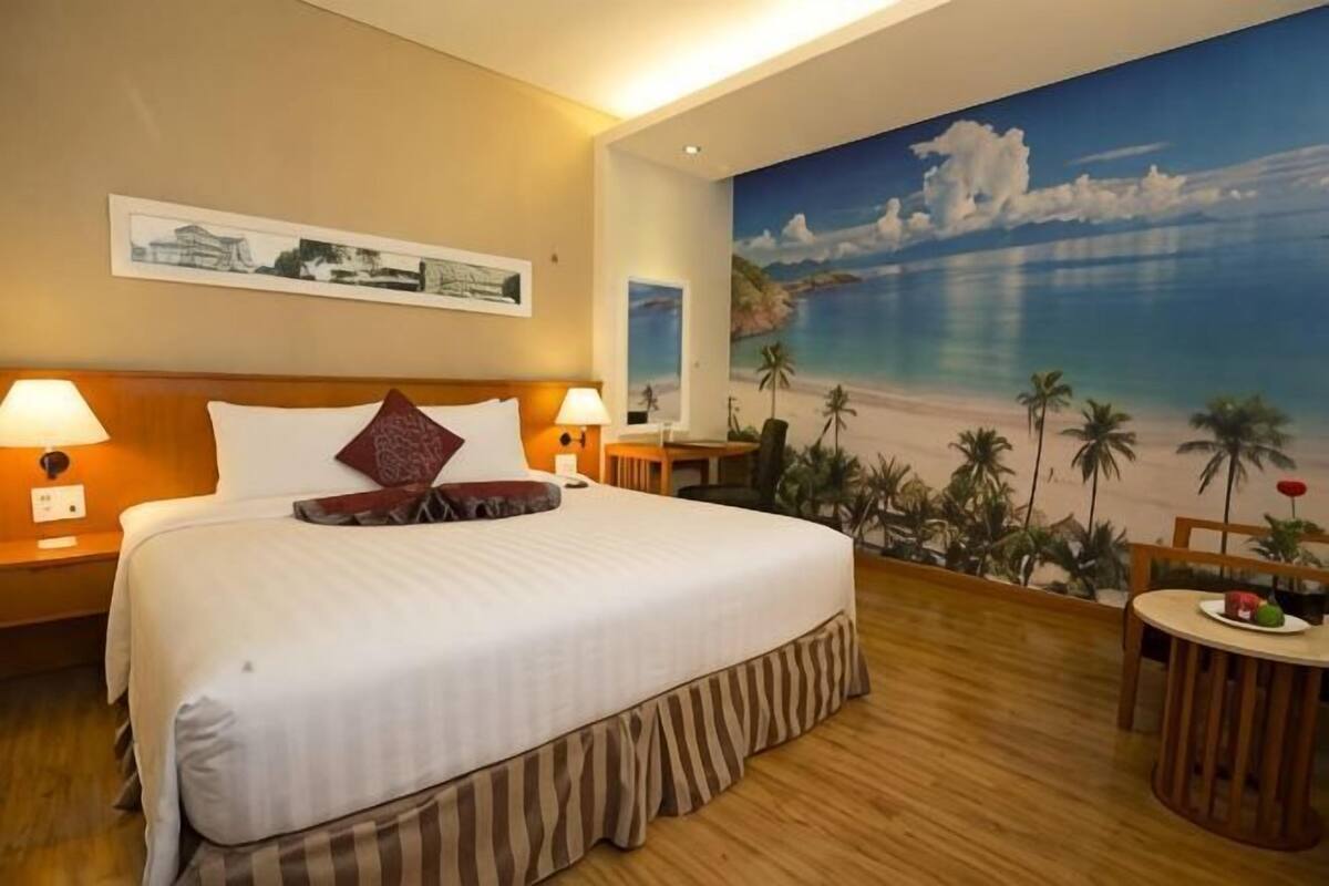 Une chambre d’hôtel avec un lit, deux tables de chevet, une petite table, un bureau, une chaise et une grande fresque représentant une plage sur le mur.