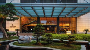 Una entrada de hotel moderna con fachada de vidrio, asientos exteriores y un jardín ajardinado.