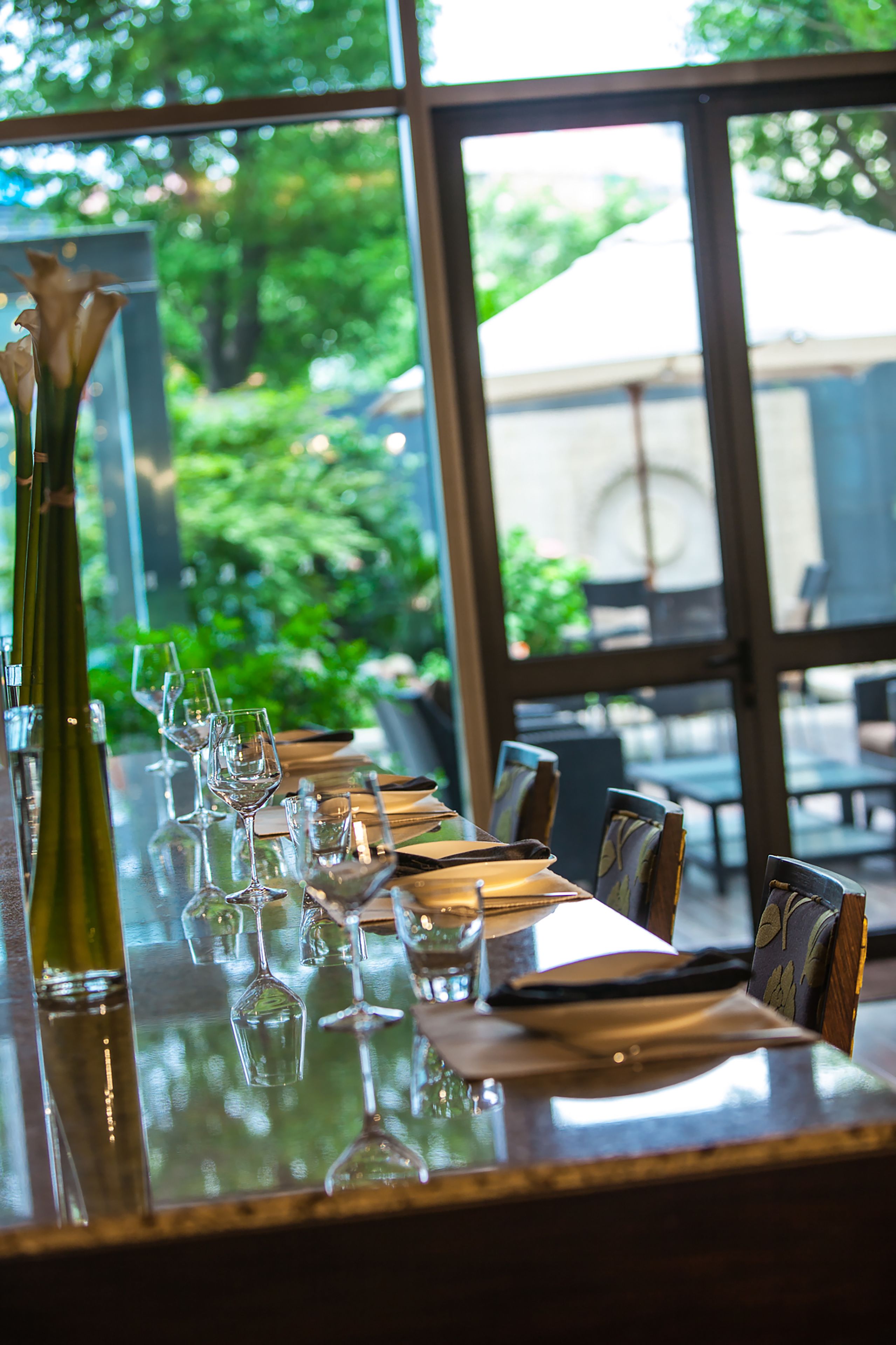 A restaurant table set with wine glasses, a vase, and cutlery, with a view of greenery outside.
