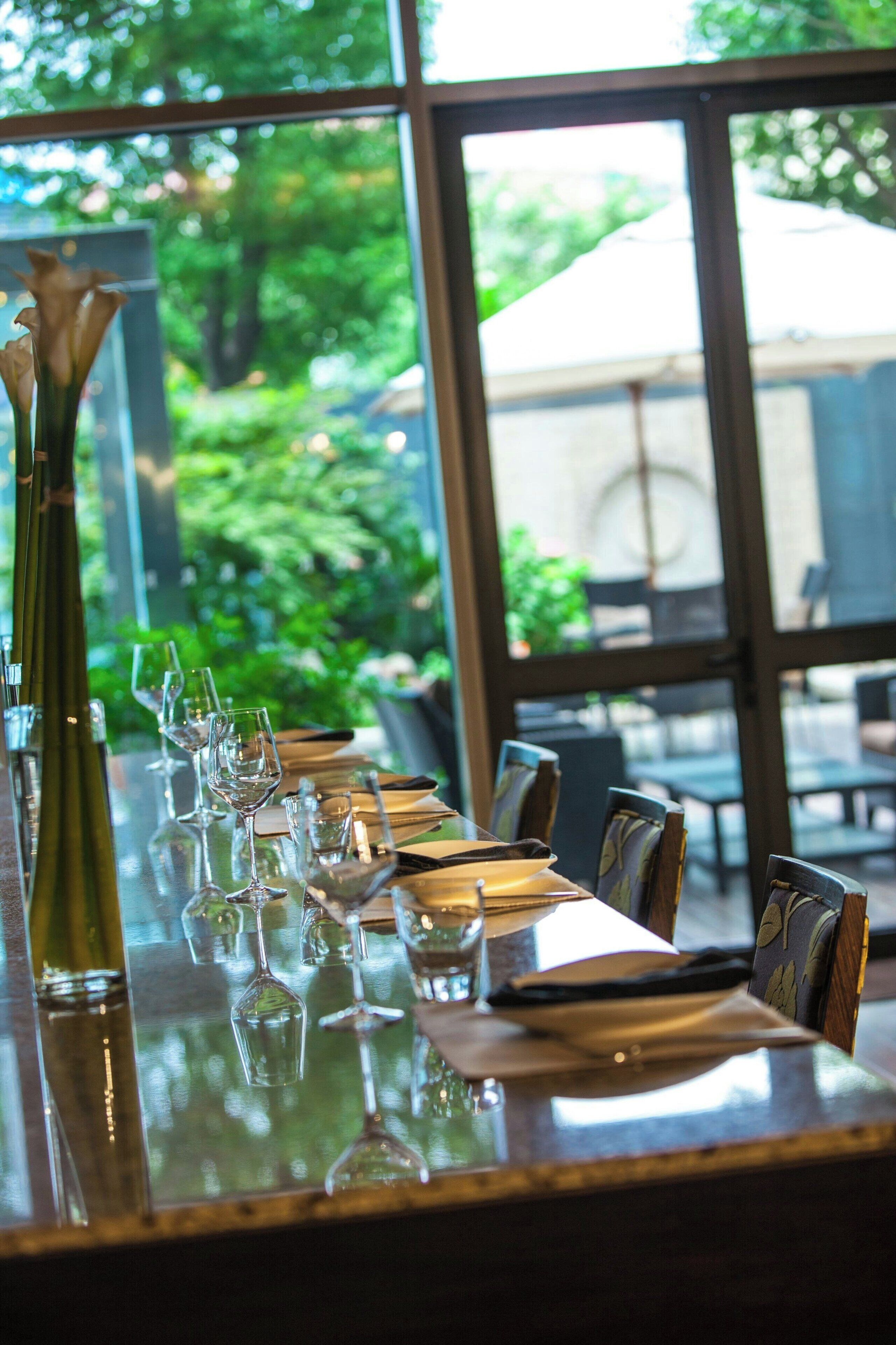 A restaurant table set with wine glasses, a vase, and cutlery, with a view of greenery outside.