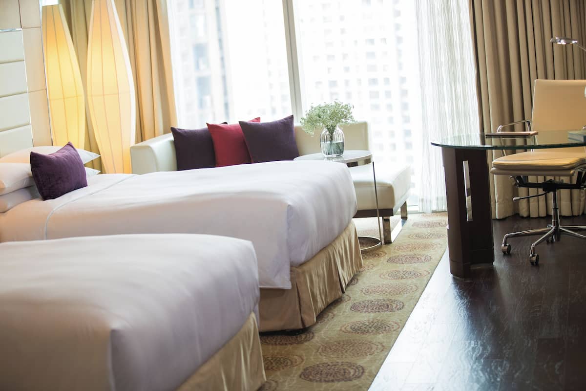 A hotel room with two beds, a desk, a chair, a lamp, and a view of the city.
