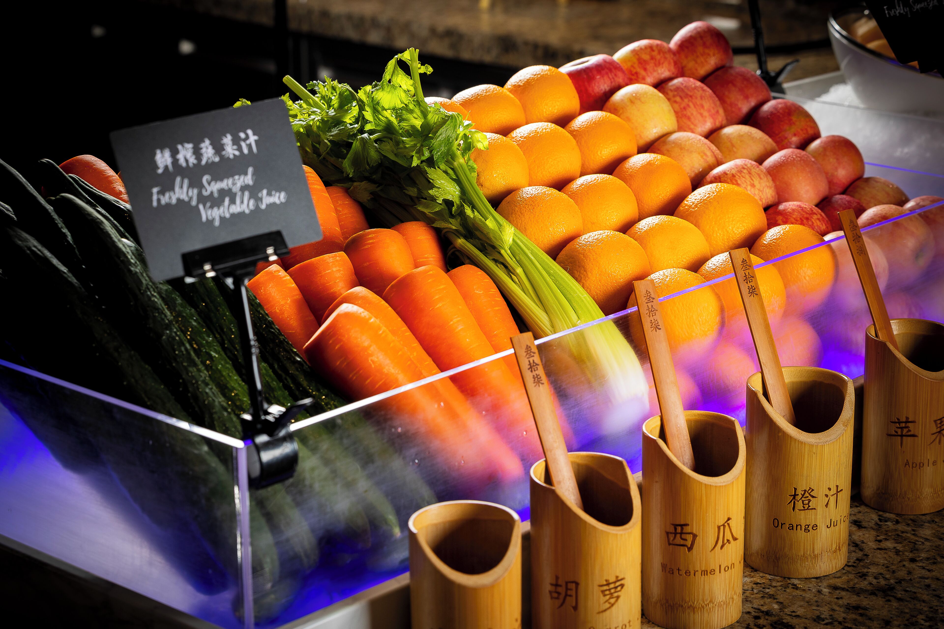 Presentatie van verse groenten en fruit, waaronder wortelen, selderij, sinaasappels en appels, met houten bakken voorzien van labels voor verschillende soorten sap.