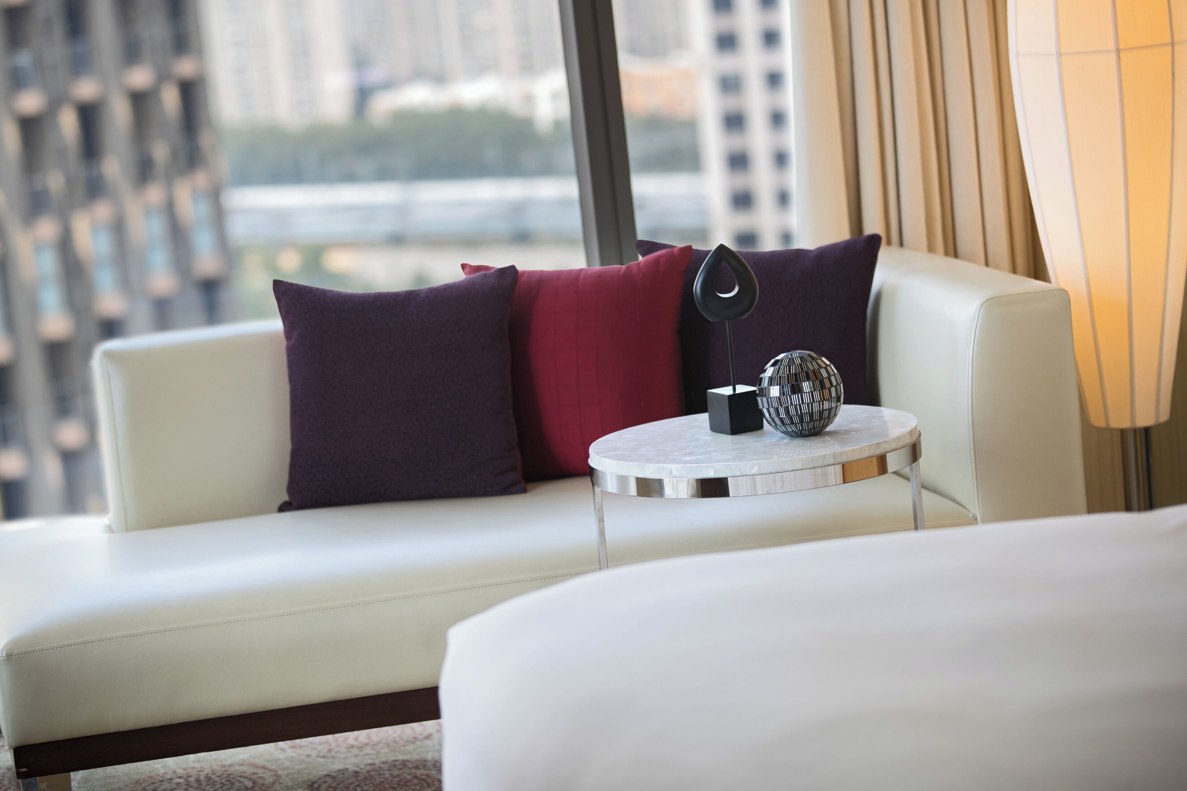 A modern hotel room with a white sofa, a round side table, and a view of a cityscape through large windows.