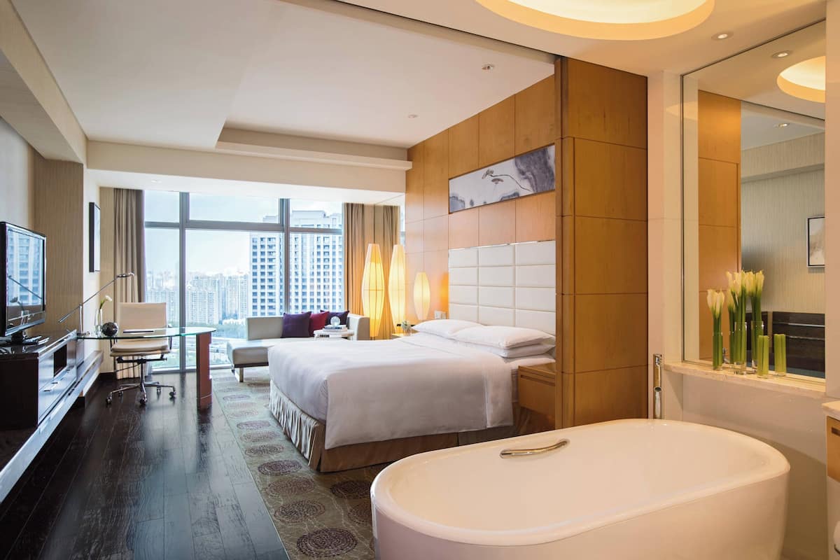 A hotel room with a large bed, a desk, a TV, a bathroom with a bathtub, and a city view.