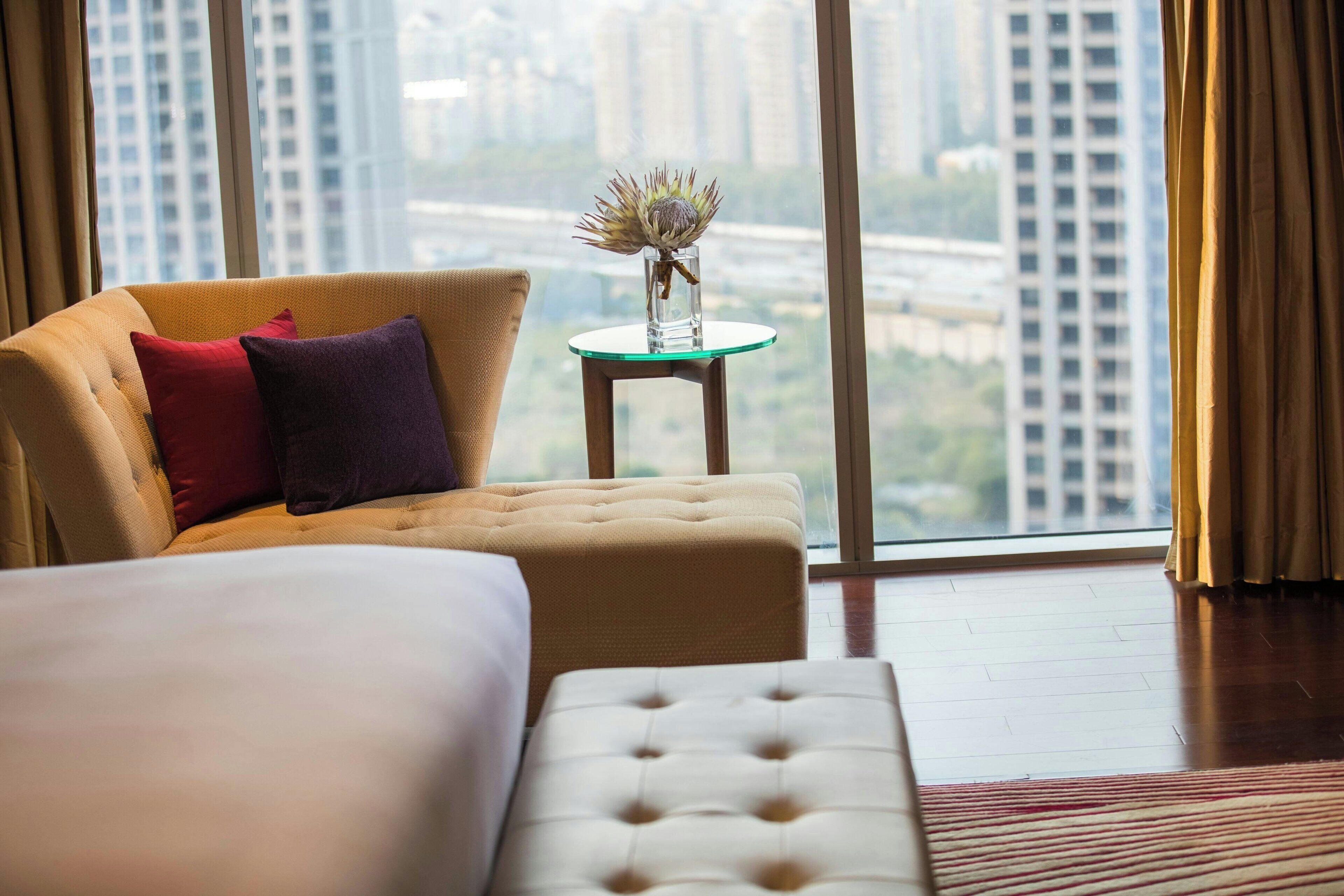 A hotel room with a bed, a sofa, a small table with a vase, and a view of tall buildings through large windows.